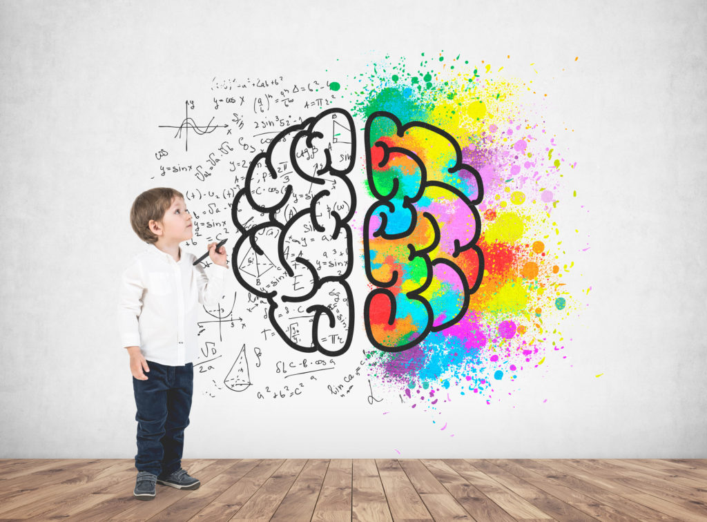 Cute little boy in a white shirt and dark jeans holding a marker and looking upwards. A concrete wall with a colorful brain sketch on it.